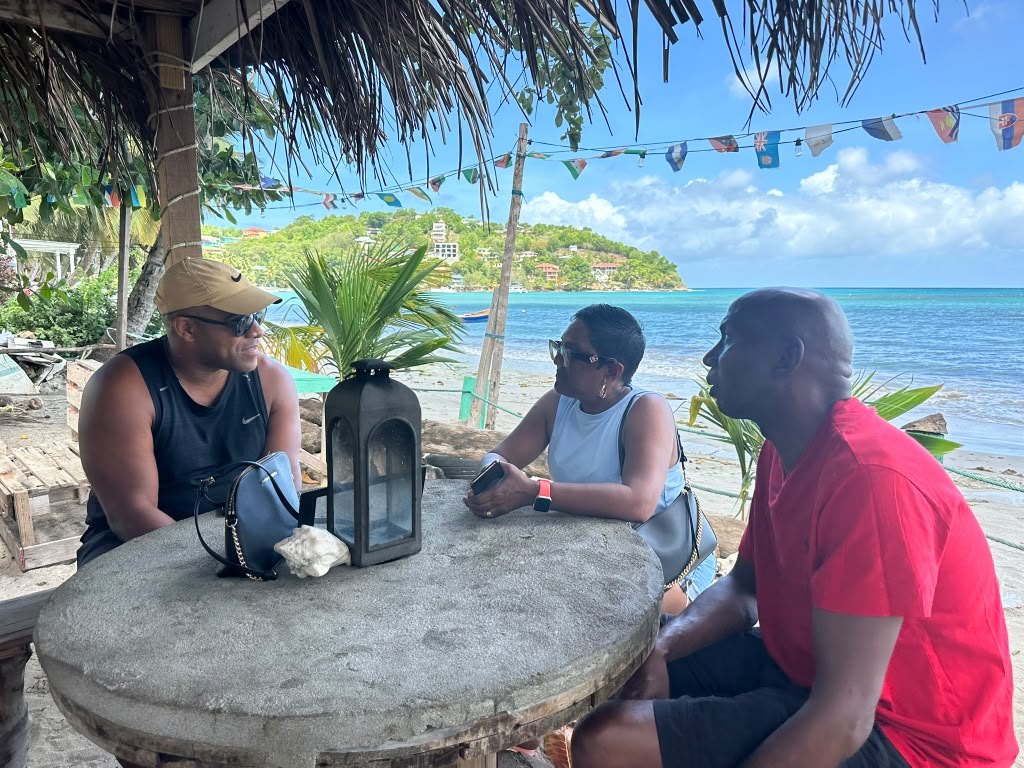Friends talking at a table at a Laborie beach bar, the sea behind them